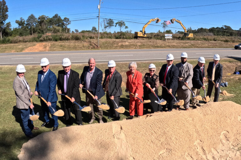 A group of officials in hard hats participate in a groundbreaking ceremony for the Chilton County Courthouse, holding shovels and standing on a dirt mound near a road. Equipment and trees form the backdrop to this pivotal moment.