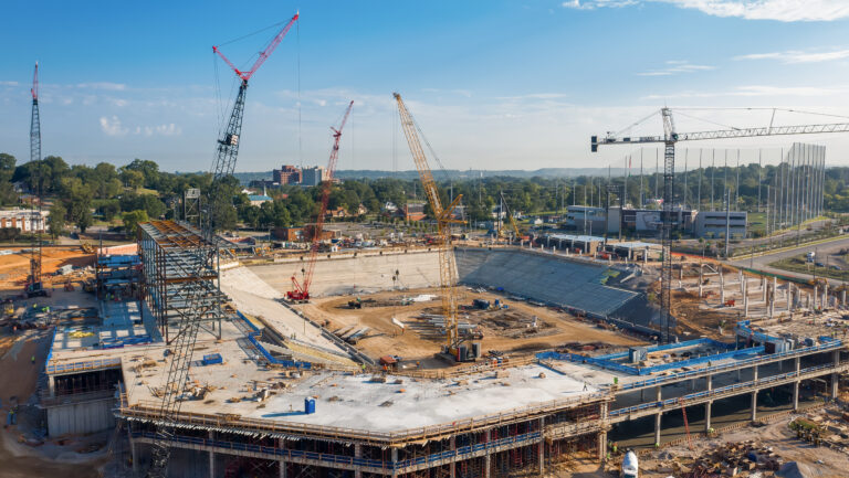 A stadium under construction, where self-perform construction methods are evident, features cranes and scaffolding amidst trees and buildings under a clear sky.