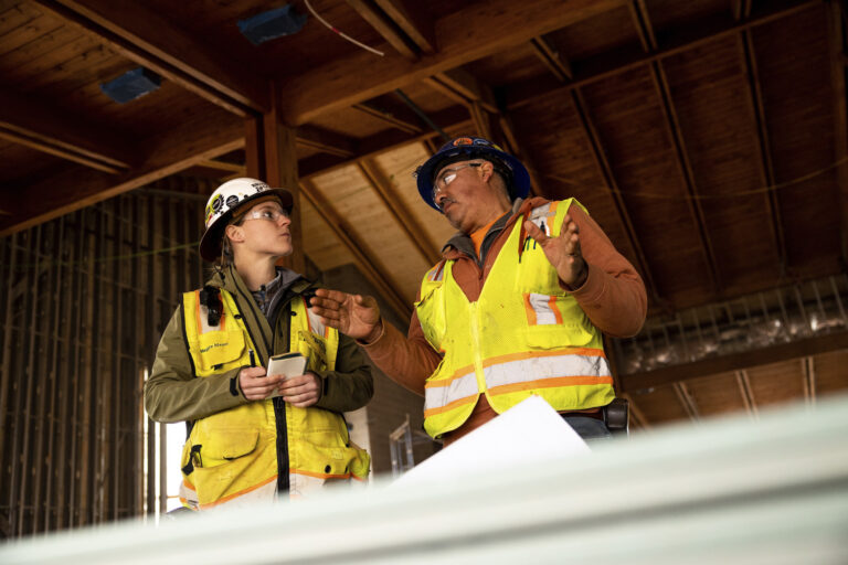 Two construction workers in safety gear and high-visibility vests discuss plans inside a wooden building under construction. One is holding a smartphone, while blueprints are partially visible.