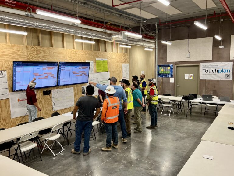 Construction workers in safety gear stand in front large screens that display project plans on a wall.