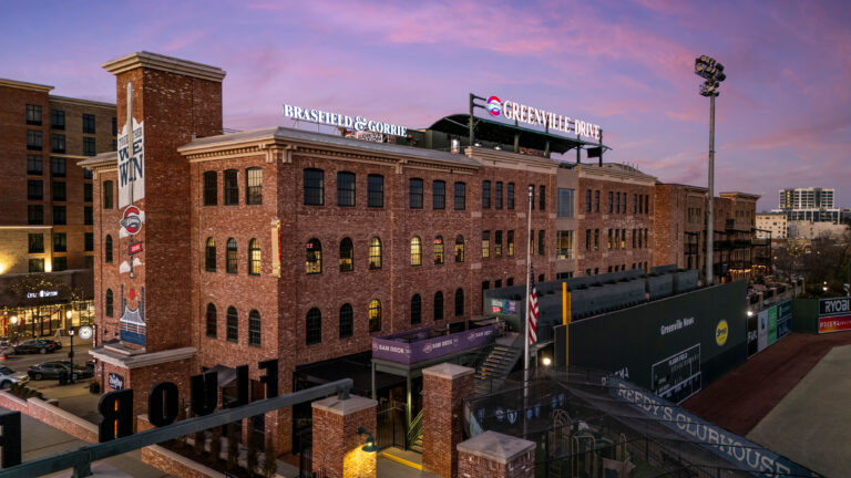 As the sun sets on the brick building, signage for Brasfield & Gorrie stands prominently. The painted mural on the side of the building adds vibrant charm, while a glimpse of a baseball field in front captures the spirit of the team’s growth at their Greenville office.