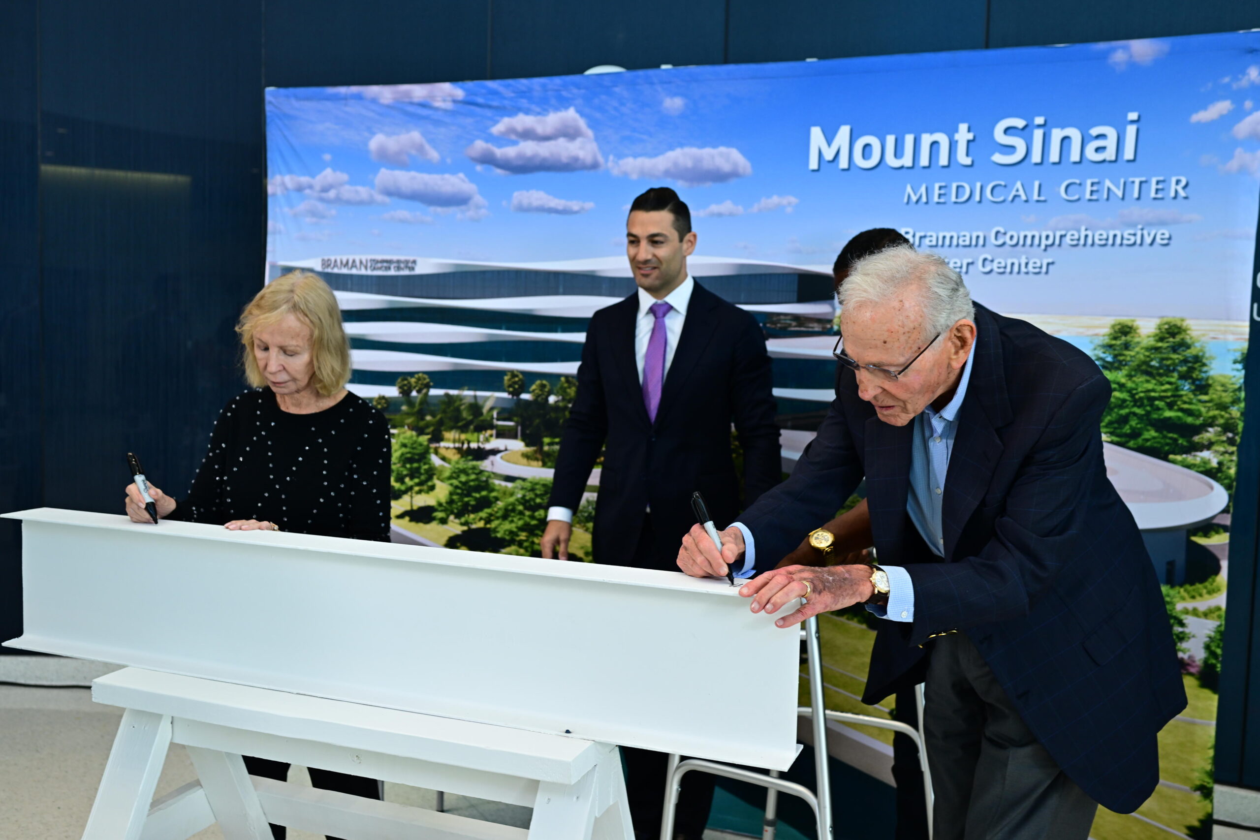 Two individuals sign a beam at a ceremony for Mount Sinai Medical Center's new structure in South Florida as a man stands behind them. A banner showcasing the center is proudly displayed in the background.