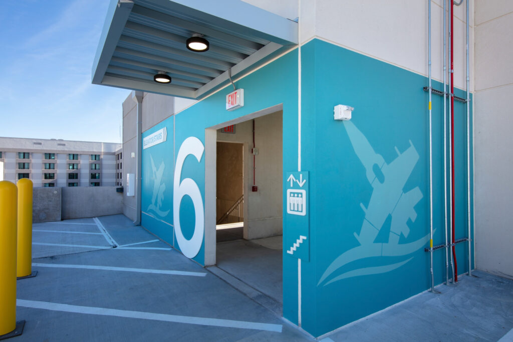 The entrance to the newly completed parking deck's elevator on floor 6. The covered area features a prominent number 6, set against blue walls and ceiling lights. Clear signage directs you to elevators and stairs, all under the bright daylight with HCA Florida West hospital building visible in the background.