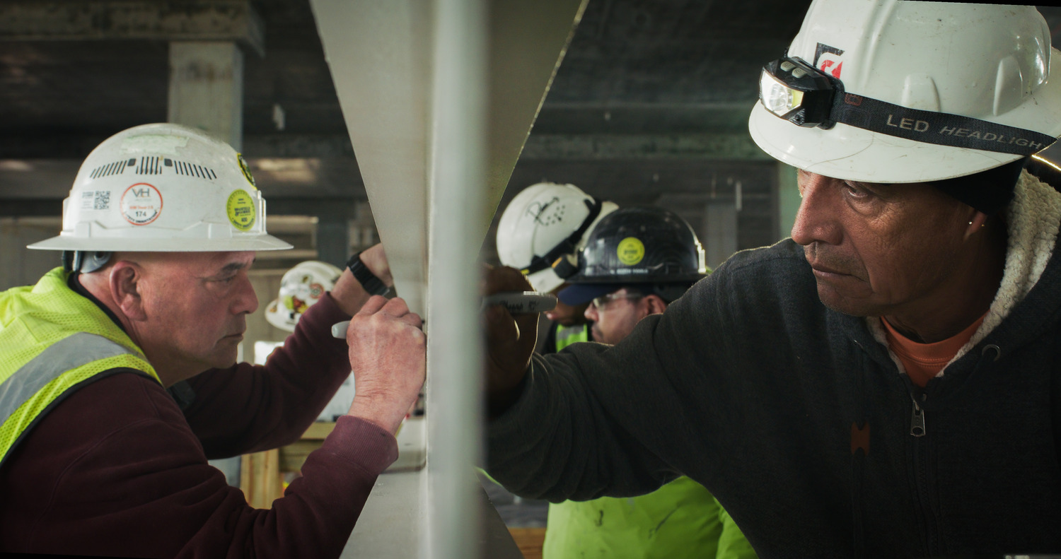 Construction workers wearing hard hats and reflective vests sign a steel beam.