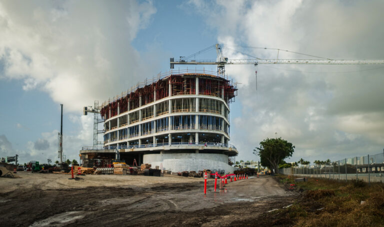 A cancer center under construction, with fluid, freeform design mimics the waves of Biscayne Bay the building will overlook. A cloudy sky looms above while a dirt path unfurls in the foreground.